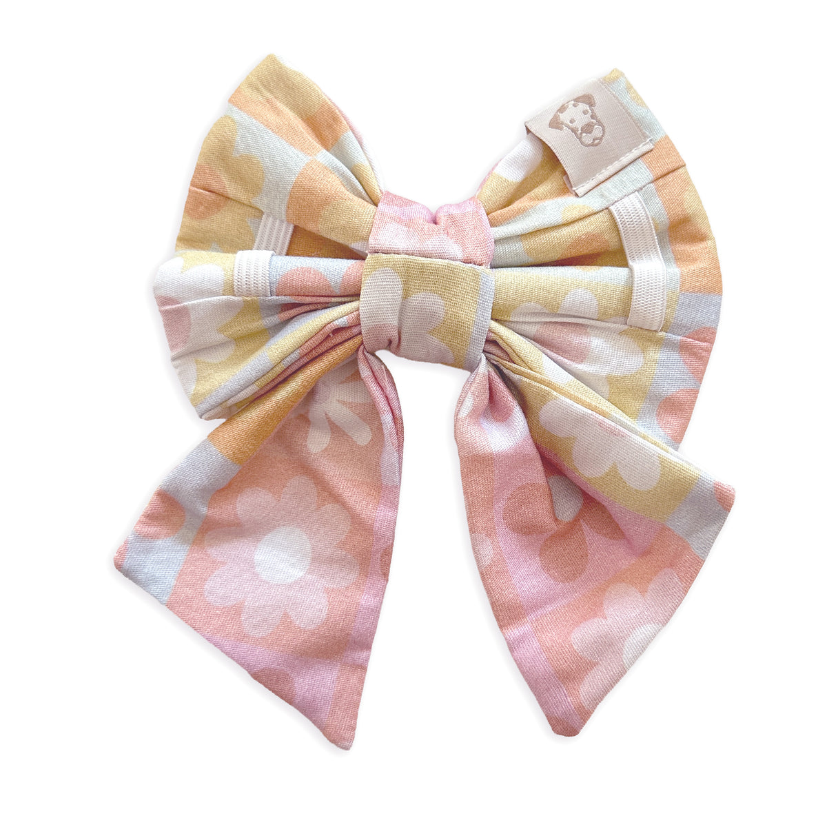 A pastel-colored fabric dog bow with a daisy pattern in pink, yellow, and light blue, featuring a small beige brand tag on the upper right side.