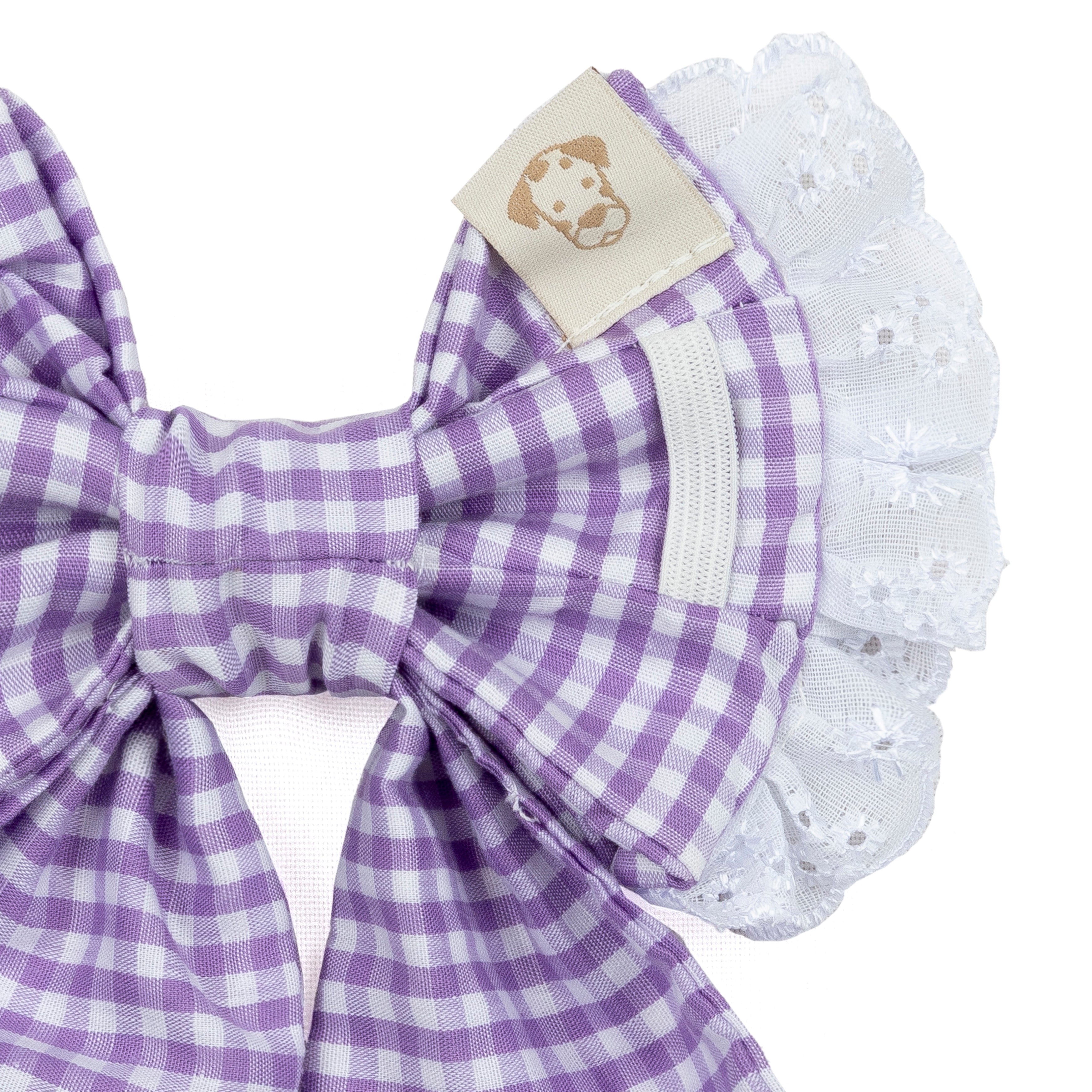 A close-up of a purple and white gingham bow accessory with a white eyelet lace ruffle and a small beige fabric tag featuring a dog silhouette.
