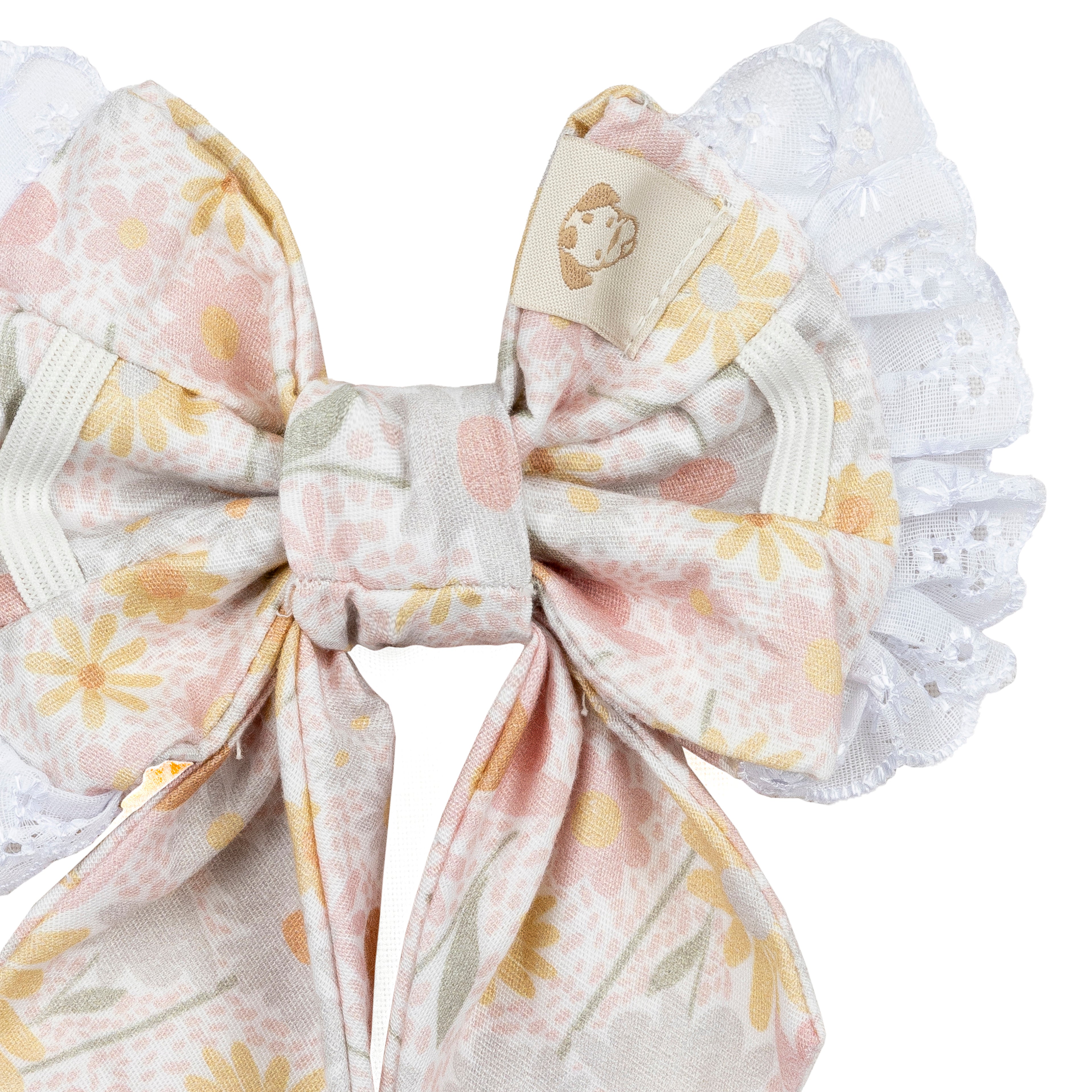 A close-up of a fabric dog bow accessory with a pastel floral print in pink, yellow, and soft green on a white base, finished with white lace ruffles and an elastic band.